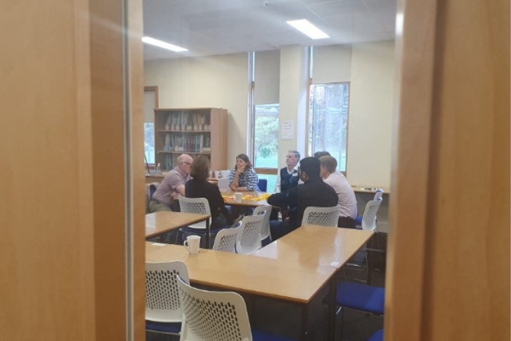 Image taken through the glass window in a door students working in small groups in a classroom style room at Homerton College