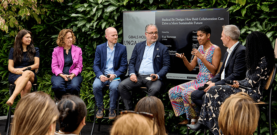 A panel of six speakers sits on stage in front of a green plant wall and an event sign. One woman speaks into a microphone while the others listen, with an audience visible in the foreground.