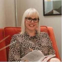 Dr Sarah Fitton is sitting in a comfortable tall-backed orange chair with a smile on her face and a book in her lap. She is looking at the camera. 