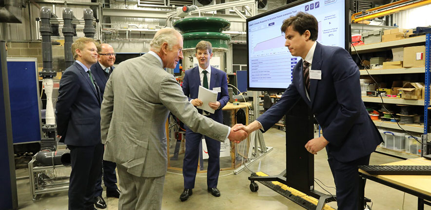 King Charles shakes hands at the Whittle Laboratory inside an industrial workshop, while several others stand nearby and a large presentation screen is visible behind them.