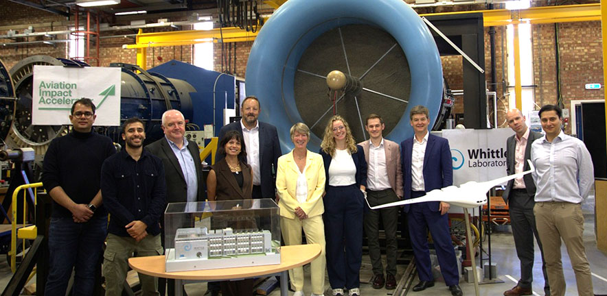 A group of people stand together in an industrial workshop in front of large turbine equipment, with display banners for the Aviation Impact Accelerator and the Whittle Laboratory visible behind them. A scale model of a facility is positioned on a table i