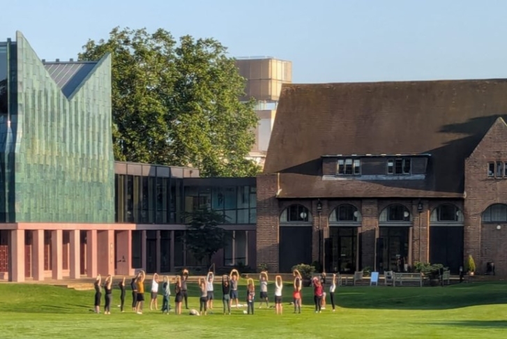 A group of students and course supervisors doing yoga on grass in front of Homerton College