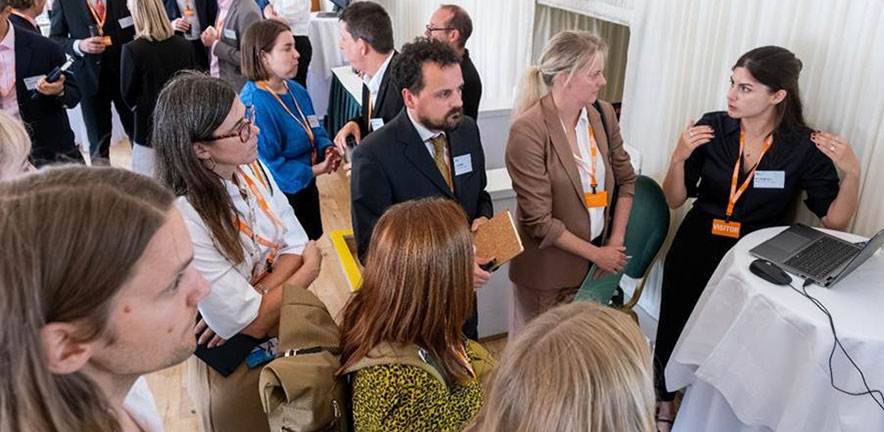 A group of people gather around a small presentation table where one person is speaking and gesturing toward a laptop screen, while others stand closely listening in a busy event setting.