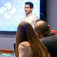 A presenter stands in front of a large screen displaying colourful charts and circular graphics, speaking to a group of people seated around a table in a meeting room.