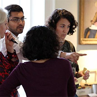 A group of four people stand together indoors, engaged in conversation. One person gestures toward something out of view while the others listen. Framed artwork hangs on the wall behind them, suggesting a professional or educational setting.