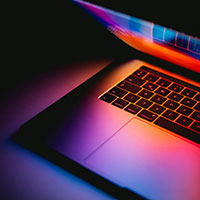 Close‑up of a laptop illuminated by colourful reflected light. A partially open laptop is shown in close‑up, with vivid pink, purple, orange, and blue light reflecting across the keyboard and lower screen. The colourful glow highlights the sleek metallic surface and creates a modern, digital aesthetic.