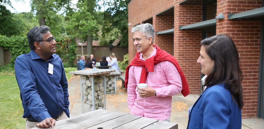 Three people talking around an outdoor wooden table in a sunny courtyard beside a brick building, with others seated and standing in the background during a social or professional gathering.