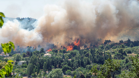 A large wildfire burns through a forested area close to several houses, with tall orange flames and thick grey smoke rising into the sky while nearby vegetation and homes sit in the foreground.