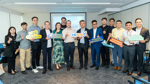 A group of thirteen people stand in a conference room giving thumbs‑up gestures and holding colourful signs with phrases such as “Life Changing,” “Europe Booster,” “Product Market Fit,” and “Pitching.” A presentation slide behind them reads “HKSTP Global Europe Booster.”