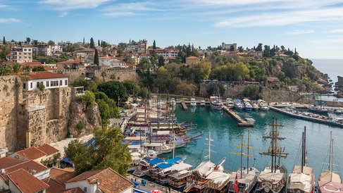 Panoramic view of Antalya’s historic harbour, with traditional boats and yachts moored in turquoise water below ancient stone fortifications. Red-roofed buildings, trees, and historic structures line the hillside above the marina, while the Mediterranean Sea stretches along the rocky coastline under a blue sky.