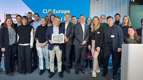 A group of people stand together in a professional setting in front of a blue backdrop displaying the text “CLG Europe” and “Corporate Leaders Group Europe.” One person in the centre holds a framed picture or certificate. The group is dressed in business or smart‑casual attire, posing for a formal event photo.