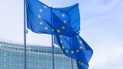 Two European Union flags fly on tall flagpoles in front of a modern curved glass building. The blue flags each display a circle of twelve yellow stars and are shown waving in the wind against the reflective façade.