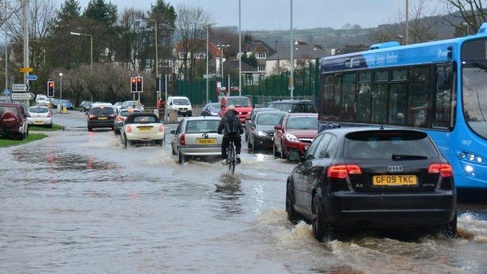 Vehicles, a bus and a cyclist travelling through a flooded urban street, illustrating the impacts of extreme weather and flood risk on transport infrastructure.
