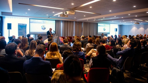 Audience seated in a modern conference room listening to a speaker at a podium, with two large presentation screens showing a portrait and text. A “Green Growth” banner stands to the left, and the room is brightly lit with ceiling lights and curtains behind the speaker.
