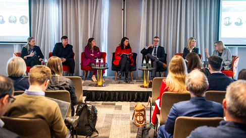 Six speakers sit on a stage in front of an audience during a professional panel discussion. They are seated on red chairs with small tables holding water bottles, while two large presentation screens behind them display slides with text and images. Attendees sit in rows, some taking notes or using laptops, in a modern conference room with grey curtains.