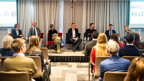 Seven speakers sit in a row on stage during a professional panel discussion, each with a small table holding water bottles and glasses. Two large presentation screens behind them display slides with headshots and text. An audience sits in rows in front of the stage, listening attentively in a modern conference room with light-coloured curtains.