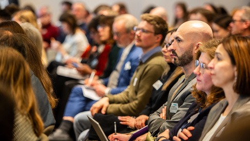 Audience members sit closely together in a conference setting, facing forward and listening attentively. Several people hold notebooks, pens, or electronic devices, suggesting they are taking notes. The group is dressed in business or business‑casual attire, creating a focused and professional atmosphere.