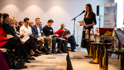A speaker stands at a podium addressing an audience seated in rows during a professional conference. Attendees listen attentively, some holding papers or taking notes. A banner to the right reads “Green Growth Summit” with University of Cambridge and CISL branding. Water bottles and glasses sit on small tables near the podium in a well‑lit meeting room.