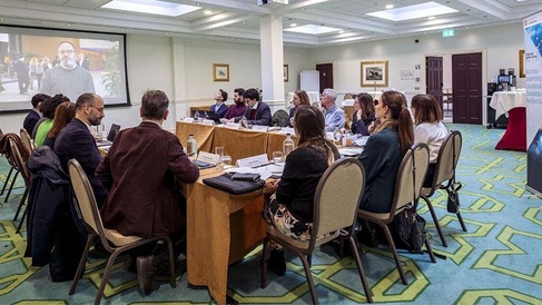 People seated around a large rectangular meeting table watch a video presentation projected on a screen at the front of the room. The video shows a person speaking outdoors. The room features patterned green carpet, framed artwork on the walls, and a banner to the right of the screen. Water glasses, papers, and nameplates are arranged on the table, indicating a formal meeting or workshop.