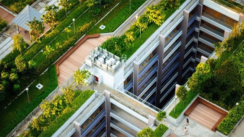 Aerial view of a modern architectural complex featuring a large rectangular courtyard surrounded by multi‑level buildings. The design includes extensive greenery such as rooftop gardens, landscaped terraces, and plant‑lined walkways. The central atrium descends several floors, with balconies and windows visible on each level.
