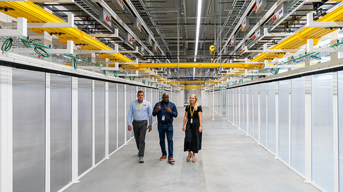 Three people walk through a brightly lit corridor at a Danfoss data centre facility, lined with rows of equipment enclosures. Overhead cable trays, power infrastructure and networking systems run the length of the facility, highlighting the scale and complexity of the digital infrastructure.