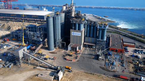 Aerial view of an industrial manufacturing facility located beside a coastal harbour. The site features large silos, processing equipment, conveyor systems, warehouses, and vehicles. Water, docks, and port infrastructure surround the facility, highlighting its connection to maritime transport and industrial operations.  Title text: Industrial manufacturing facil