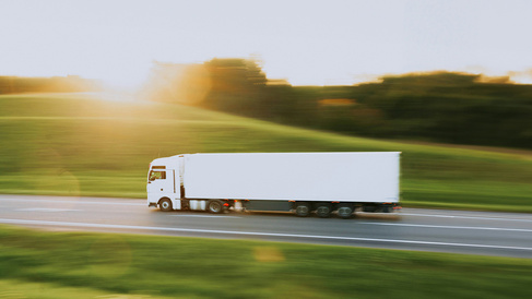 White articulated lorry travelling along a motorway through a rural landscape at sunset. Motion blur in the surrounding fields and trees emphasises the vehicle's movement, while warm sunlight illuminates the scene.