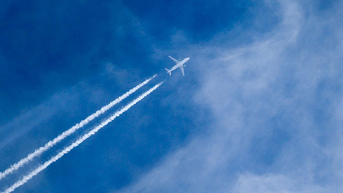 A white airplane flying high in a blue sky, leaving two long white contrails behind it as it moves through thin, wispy clouds.