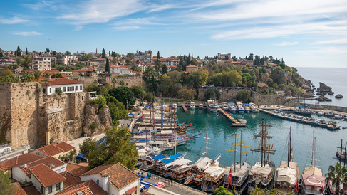 Panoramic view of Antalya’s historic harbour, with traditional boats and yachts moored in turquoise water below ancient stone fortifications. Red-roofed buildings, trees, and historic structures line the hillside above the marina, while the Mediterranean Sea stretches along the rocky coastline under a blue sky.