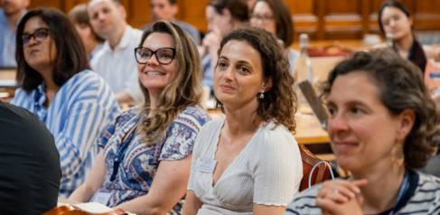 Four female students smiling and watching a presentation with other students in the background. 