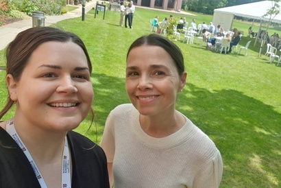 Emma Fromberg and Simona Azzolini stood smiling close together with a group of students sat around round plastic tables in front of a white marquee