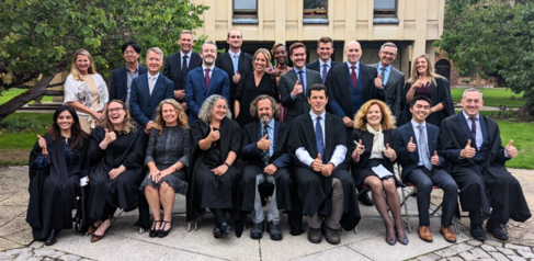 image of 22 people wearing black formal graduation robes holding their thumbs up and smiling