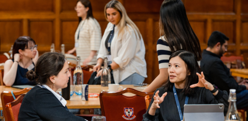 Two female students sat at a table in the foreground, one making gestures with her hands and smiling. Four women and one man are in the background sat and stood around other tables. 
