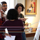 A group of four people stand around a flip chart in a bright meeting room. One person gestures toward the chart while another holds a marker and speaks, and the others watch with papers and sticky notes in hand. A framed portrait, a lamp, and a bowl of fruit sit in the background, giving the scene a collaborative workshop atmosphere.