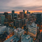 Aerial view of a modern city at sunset, with tall illuminated skyscrapers, dense urban streets below, and mountains in the distance under an orange‑pink sky.