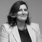 A black and white photo of Marija, in a smart blazer, dark top and thick necklace. Marija is smiling at the camera and looks professional. 