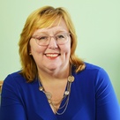 Jennifer, a white woman with light ginger hair, glasses, and a layered necklace is smiling at the camera in a blue dress.