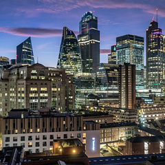 Twilight view of central London’s skyline, featuring The Gherkin, The Leadenhall Building, and other illuminated skyscrapers rising above older brick buildings under a blue‑purple dusk sky.
