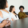 Five professionals sit around a large conference table in a modern meeting room, engaged in discussion while one person speaks and the others listen attentively.