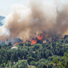 A large wildfire burns through a forested area close to several houses, with tall orange flames and thick grey smoke rising into the sky while nearby vegetation and homes sit in the foreground.