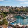 Panoramic view of Antalya’s historic harbour, with traditional boats and yachts moored in turquoise water below ancient stone fortifications. Red-roofed buildings, trees, and historic structures line the hillside above the marina, while the Mediterranean Sea stretches along the rocky coastline under a blue sky.