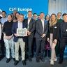 A group of people stand together in a professional setting in front of a blue backdrop displaying the text “CLG Europe” and “Corporate Leaders Group Europe.” One person in the centre holds a framed picture or certificate. The group is dressed in business or smart‑casual attire, posing for a formal event photo.
