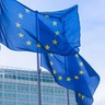 Two European Union flags fly on tall flagpoles in front of a modern curved glass building. The blue flags each display a circle of twelve yellow stars and are shown waving in the wind against the reflective façade.