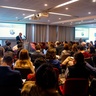 Audience seated in a modern conference room listening to a speaker at a podium, with two large presentation screens showing a portrait and text. A “Green Growth” banner stands to the left, and the room is brightly lit with ceiling lights and curtains behind the speaker.