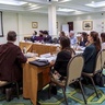 People seated around a large rectangular meeting table watch a video presentation projected on a screen at the front of the room. The video shows a person speaking outdoors. The room has patterned green carpet, framed artwork on the walls, and a banner to the right of the screen. Water glasses, papers, and nameplates are arranged on the table, indicating a formal meeting or workshop.