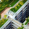 Aerial view of a modern architectural complex featuring a large rectangular courtyard surrounded by multi‑level buildings. The design includes extensive greenery such as rooftop gardens, landscaped terraces, and plant‑lined walkways. The central atrium descends several floors, with balconies and windows visible on each level.
