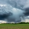 Dark storm clouds over an agricultural field with heavy rainfall visible in the distance beneath a dramatic thunderstorm sky.