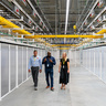 Three people walk through a brightly lit corridor at a Danfoss data centre facility, lined with rows of equipment enclosures. Overhead cable trays, power infrastructure and networking systems run the length of the facility, highlighting the scale and complexity of the digital infrastructure.