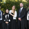Five men and two women smiling and stood in a line wearing University of Cambridge robes over formal wear.  