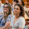 Four female students smiling and watching a presentation with other students in the background. 
